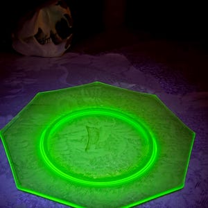 May include: A glowing green octagonal glass plate with a floral pattern. The plate is illuminated, showcasing its design. A skull is visible in the background, adding a contrasting element to the composition.
