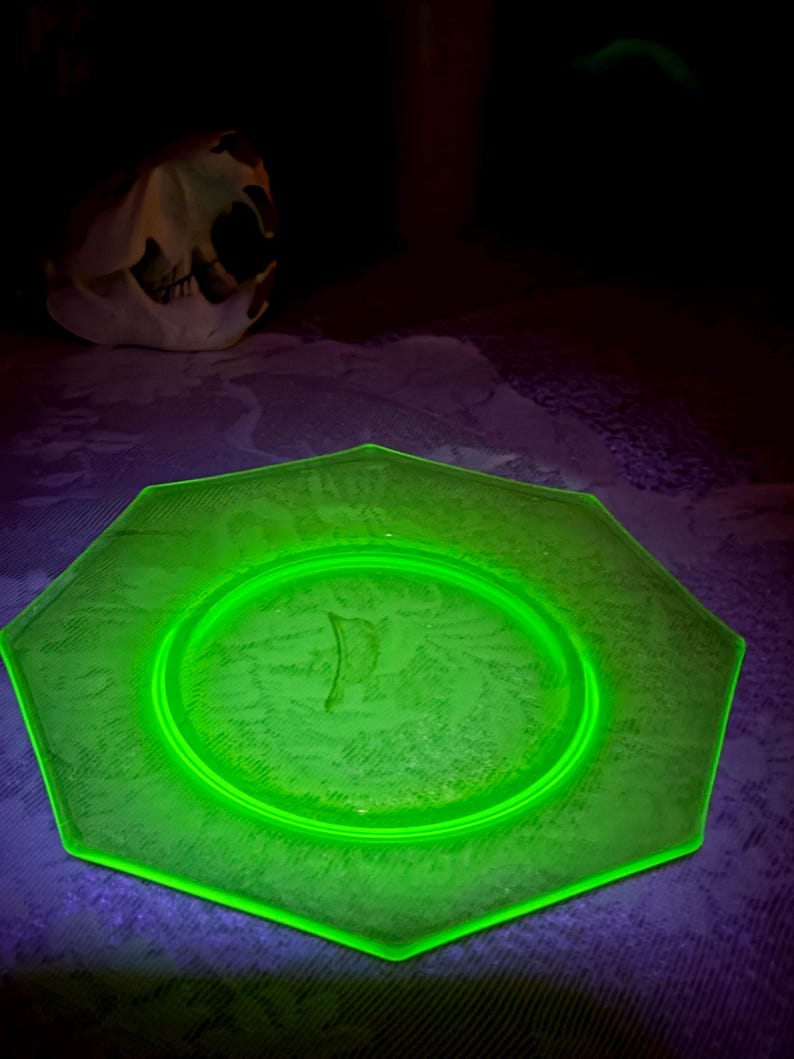 May include: A glowing green octagonal glass plate with a floral pattern. The plate is illuminated, showcasing its design. A skull is visible in the background, adding a contrasting element to the composition.