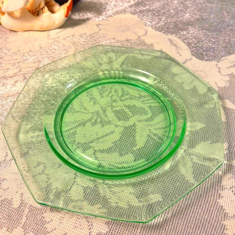 May include: A vintage, octagonal, light green glass plate. The plate has a circular center and a slightly textured surface. The plate is set on a floral patterned tablecloth. The plate is a piece of glassware.
