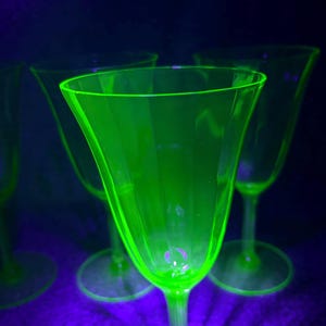 Può includere: Un set di quattro calici in vetro tinto di verde. I bicchieri hanno una coppa a forma di tulipano e un gambo sottile, con una base circolare. I bicchieri sono illuminati da una brillante luce verde su sfondo scuro.