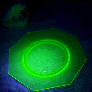 May include: A glowing green octagonal glass plate with a floral pattern. The plate has a raised rim and a central depression. The plate is illuminated with a black light, causing it to glow brightly.