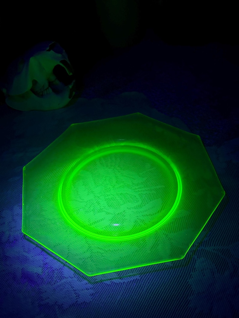May include: A glowing green octagonal glass plate with a floral pattern. The plate has a raised rim and a central depression. The plate is illuminated with a black light, causing it to glow brightly.