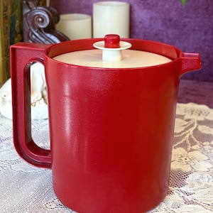 May include: A vintage red plastic pitcher with a white lid and handle. The pitcher has a textured surface and a classic design. The lid features a red knob. This retro kitchenware item is perfect for serving beverages.