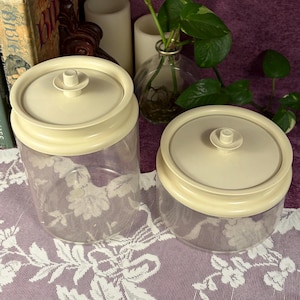 May include: Two clear glass canisters with off-white plastic lids. Each lid features a small, centered circular knob for easy lifting. The canisters have a cylindrical shape with a smooth, transparent body, providing a clear view of the contents inside.