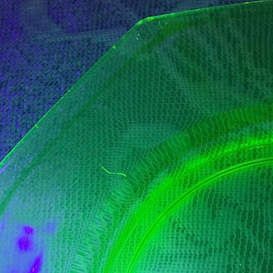 May include: A close-up of a green glass plate with a curved edge. The plate has a textured surface and a bright neon green glow. The background is a dark blue and green fabric.