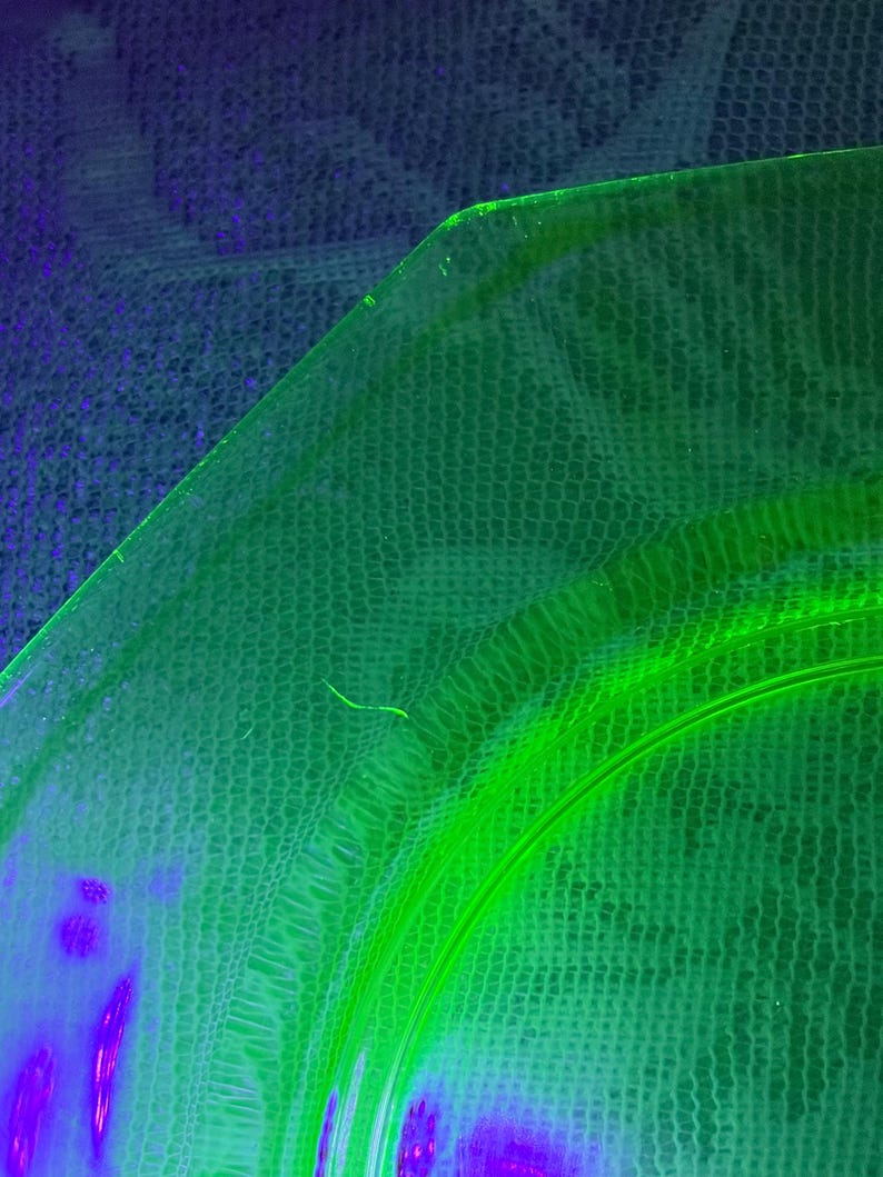 May include: A close-up of a green glass plate with a curved edge. The plate has a textured surface and a bright neon green glow. The background is a dark blue and green fabric.