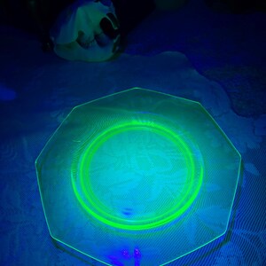 May include: A hexagonal, clear glass plate with a bright green rim and center circle. The plate has a floral pattern in the center. The image is lit with a black light, causing the glass to glow. A white object is in the background.