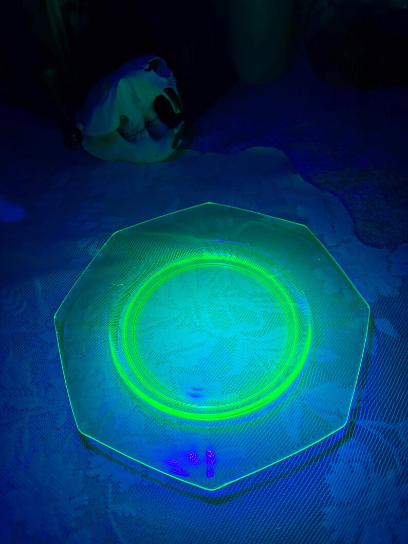 May include: A hexagonal, clear glass plate with a bright green rim and center circle. The plate has a floral pattern in the center. The image is lit with a black light, causing the glass to glow. A white object is in the background.
