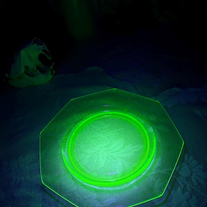 May include: A glowing green octagonal glass plate with a circular design in the center. The plate is illuminated, showcasing its edges and the pattern within. Other green glass objects are in the background.