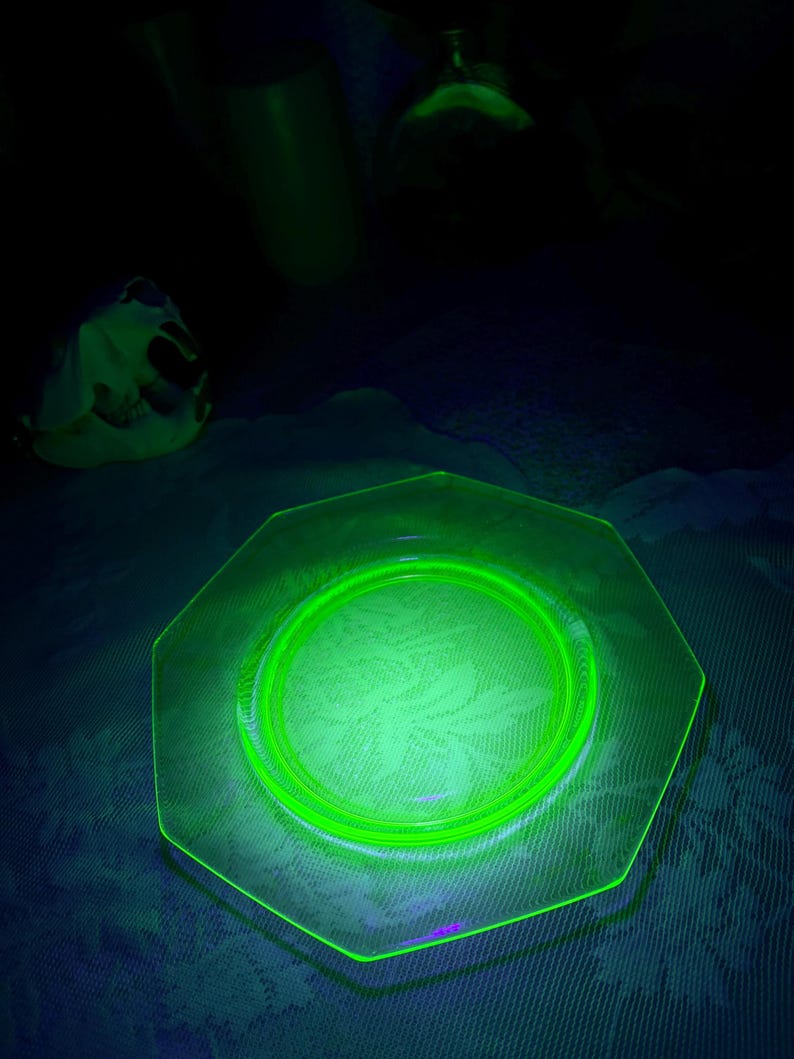 May include: A glowing green octagonal glass plate with a circular design in the center. The plate is illuminated, showcasing its edges and the pattern within. Other green glass objects are in the background.