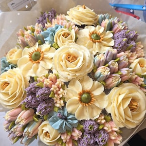 Könnte beinhalten: Ein Blumenstrauß aus Buttercreme mit gelben, hellblauen, rosa und lila Blüten. Die Anordnung umfasst Rosen, Gänseblümchen und andere Blütenformen, verpackt in braunem Papier.