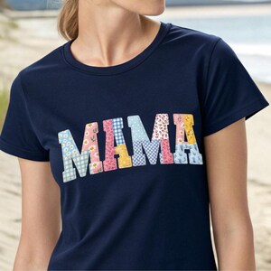Puede incluir: Camiseta azul marino con la palabra "MAMA" en letras de patchwork coloridas. Las letras presentan patrones florales, de cuadros y lisos en tonos de azul, rosa, amarillo y blanco. La camiseta tiene cuello redondo y mangas cortas.
