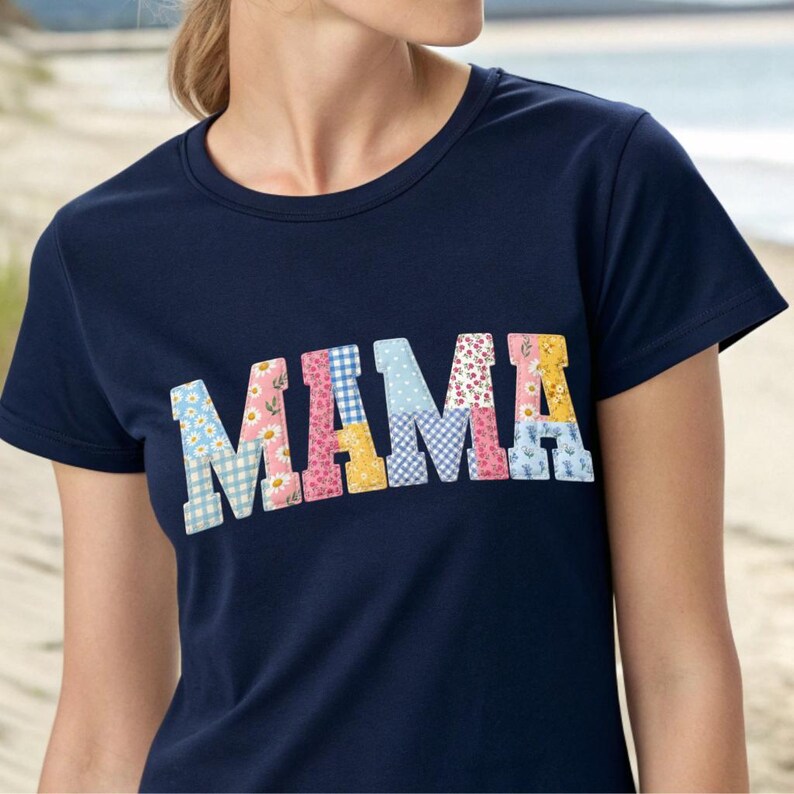 Puede incluir: Camiseta azul marino con la palabra "MAMA" en letras de patchwork coloridas. Las letras presentan patrones florales, de cuadros y lisos en tonos de azul, rosa, amarillo y blanco. La camiseta tiene cuello redondo y mangas cortas.