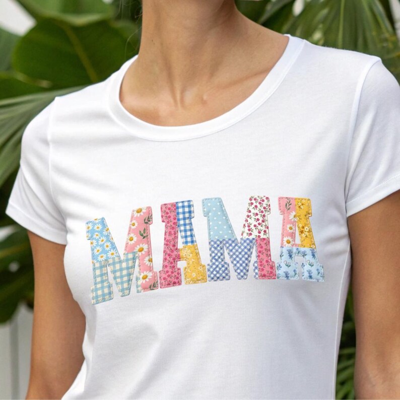 Puede incluir: Camiseta blanca con la palabra "MAMA" en letras de patchwork coloridas. Las letras presentan patrones florales, de cuadros y de lunares en tonos de azul, rosa y amarillo. La camiseta tiene cuello redondo.