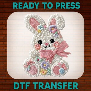 May include: A white bunny-shaped DTF transfer with a textured surface, decorated with embroidered flowers and a pink bow. The design includes the text "READY TO PRESS" at the top and "DTF TRANSFER" at the bottom.