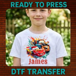 May include: White t-shirt with a vibrant graphic of two cartoon cars, and the name "James" in red script. The shirt has the text "READY TO PRESS" and "DTF TRANSFER" in teal.