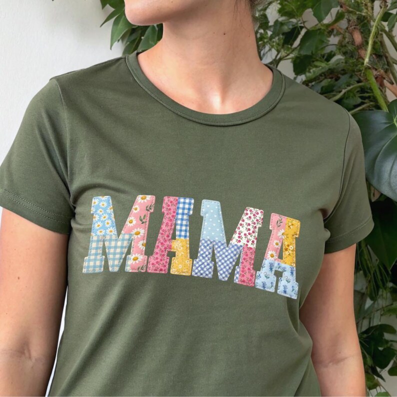 Puede incluir: Camiseta verde oliva con la palabra "MAMA" en letras de patchwork coloridas. Las letras presentan patrones florales, de cuadros y de lunares en tonos azules, rosas y amarillos. La camiseta tiene cuello redondo y mangas cortas.