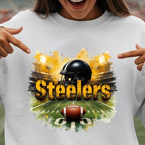 Pu&ograve; includere: Felpa bianca con un design da campo da football, un casco nero e un pallone da football. La parola "Steelers" &egrave; in lettere dorate. Lo sfondo include luci dello stadio e una combinazione di colori giallo e verde.
