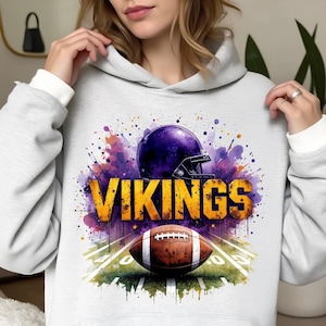 Può includere: Una felpa con cappuccio grigio chiaro con un casco da football americano viola, un pallone da football marrone e la scritta "VIKINGS" in lettere dorate. Il design include schizzi di vernice colorati e un campo verde.