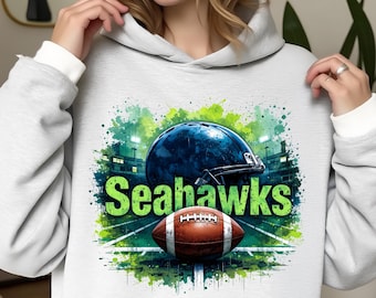 Könnte beinhalten: Hellgrauer Kapuzenpullover mit einem Grafikdesign eines Footballhelms und -balls mit dem Wort "Seahawks" in Grün. Das Design hat einen Aquarelleffekt mit grünen und blauen Akzenten.
