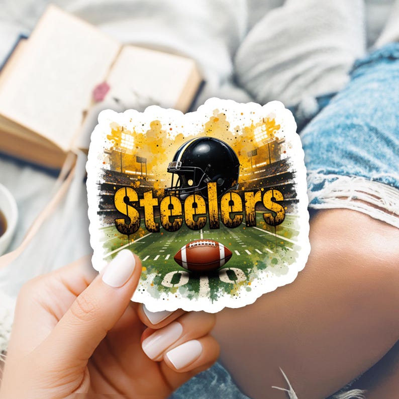Pu&ograve; includere: Un adesivo raffigurante una scena di stadio di football con la scritta "Steelers" in giallo e nero. Sono visibili anche un casco da football nero e un pallone da football marrone e bianco. L'adesivo ha un bordo bianco.