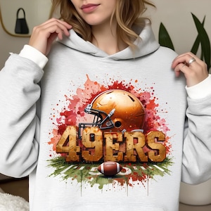 Può includere: Una felpa con cappuccio grigio chiaro con un design ad acquerello di un casco da football, un pallone da football e la scritta "49ERS" in un carattere strutturato e invecchiato. Il design include schizzi di vernice rossa e arancione ed erba verde.