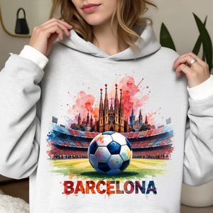 May include: A light grey hoodie with a watercolour design of a football on a green pitch, in front of a stadium and the Sagrada Família. The word "BARCELONA" is printed in colourful letters below.