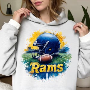 Puede incluir: Una sudadera con capucha blanca con un diseño de acuarela de un casco de fútbol americano azul, un balón de fútbol marrón y la palabra "Rams" en amarillo. El diseño incluye un campo verde y salpicaduras de pintura amarilla y azul.