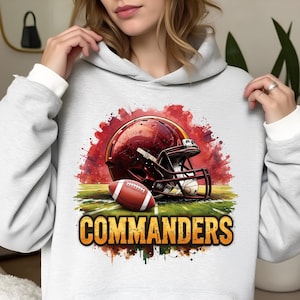 May include: A light gray hoodie featuring a football helmet and football with the word "COMMANDERS" in gold. The helmet is maroon and gold, and the football is brown and white. The design is set against a red and green background.