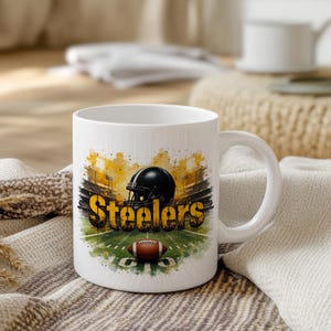 Pu&ograve; includere: Tazza in ceramica bianca con un design a tema football. Il design include un casco nero, la scritta "Steelers" in oro e un pallone da football marrone su un campo verde. La tazza ha un manico bianco.