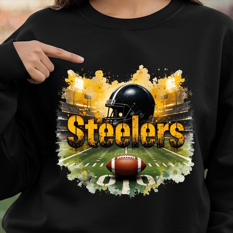Pu&ograve; includere: Felpa nera con una grafica. Il design presenta un casco da football nero, un pallone da football marrone e la scritta "Steelers" in giallo e oro. Lo sfondo &egrave; uno stadio con luci gialle.