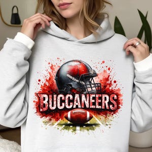 Puede incluir: Sudadera gris claro con un diseño de fútbol. El diseño presenta un casco de fútbol negro y rojo, un balón de fútbol marrón y la palabra "BUCCANEERS" en blanco. El diseño tiene un efecto acuarela con salpicaduras rojas.