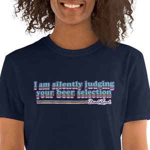 May include: A navy blue t-shirt with the text "I am silently judging your beer selection" in a retro font with colourful stripes. The text "Drink Local" is printed below the main text.