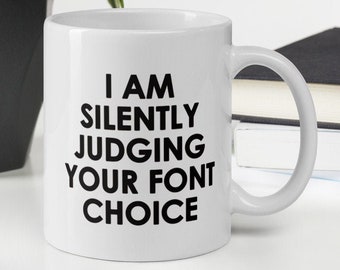 Puede incluir: Taza de cerámica blanca con el texto "I AM SILENTLY JUDGING YOUR FONT CHOICE" en negro. La taza tiene un asa curva y está sobre una superficie blanca. Un teclado, una planta y libros están en el fondo.