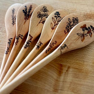 May include: Six wooden spoons with botanical illustrations and herb names: rosemary, thyme, parsley, sage, oregano, and basil. The spoons are arranged on a wooden surface, showcasing their natural wood grain and engraved details.