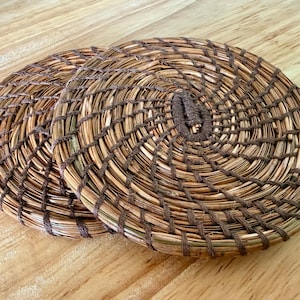 Pode incluir: Duas bases para copos redondas, tecidas com palha de cor natural e barbante marrom escuro. As bases para copos têm um design em espiral e são colocadas sobre uma superfície de madeira marrom clara. Cada base para copos tem aproximadamente 10 cm de diâmetro.