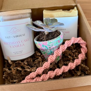 May include: A gift box containing a vanilla-scented candle, a small succulent in a floral pot, a bar of soap, and a pink macrame bracelet. The candle label reads "Salt + Sawdust Birthday Cake Vanilla". The box is filled with brown paper filler.