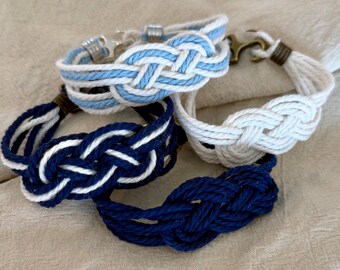 Op de afbeelding: Vier nautische armbanden in blauw en wit. De armbanden zijn gemaakt van gevlochten touw, met een knoopontwerp. Eén armband is effen marineblauw, een andere is wit en twee zijn blauw en wit.