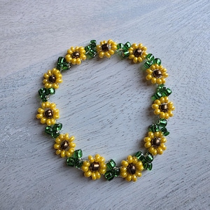 May include: A beaded bracelet featuring a sunflower design. The bracelet has yellow and brown flower beads with green leaf accents. The bracelet is made of small, round beads strung together to form the floral pattern.