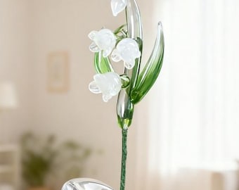 May include: A clear glass lily of the valley flower sculpture with white blossoms and green leaves. The flower is displayed in a clear glass vase. The stem is a dark green color.