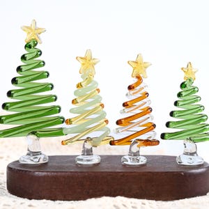 Peut inclure: Quatre décorations de Noël en verre en vert, jaune et orange, chacune surmontée d'une étoile jaune. Les arbres sont montés sur une base en bois brun foncé, créant un affichage festif.