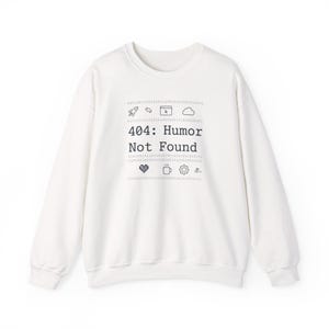 Puede incluir: Sudadera blanca con el texto negro "404: Humor Not Found", acompañado de pequeños iconos gráficos. El diseño incluye un cohete, una carpeta, una nube, un corazón, una taza de café y un engranaje.