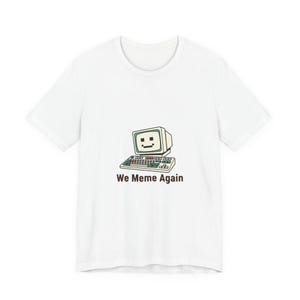 May include: White t-shirt featuring a retro computer graphic with a smiling face on the screen and a keyboard. The text "We Meme Again" is printed below the graphic in a bold, black font.