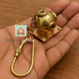 May include: A brass-colored keychain featuring a miniature diving helmet design. The helmet is attached to a square base and a chain, with a clasp for attaching keys. The keychain is held in a hand.
