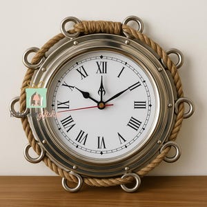 Könnte beinhalten: Eine runde Wanduhr im nautischen Stil mit einem silberfarbenen Metallrahmen und einem weißen Zifferblatt mit schwarzen römischen Ziffern. Die Uhr ist von einem dicken, braunen Seil und Metallringen umgeben. Die Zeiger sind schwarz.