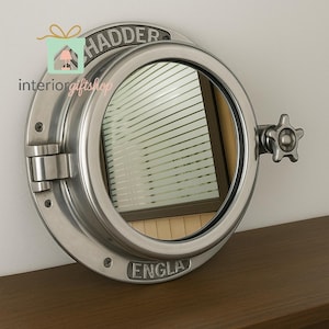 May include: A round, silver-colored porthole mirror with the word "HADDER" at the top and "ENGLA" at the bottom. The mirror has a star-shaped handle and is mounted on a brown surface.