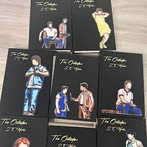 May include: A collection of eight black books titled The Outsiders by S.E. Hinton. Each cover features a unique, colorful, stylized illustration of a character from the story, with the title and author name printed in gold lettering at the top.