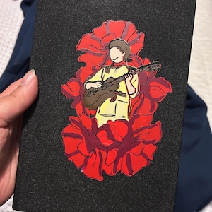 May include: Black journal with a red and yellow illustration of a person playing a guitar inside a red flower. The journal has a textured cover and a ribbon bookmark.