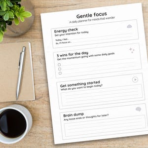 May include: A daily planner titled "Gentle Focus" with sections for daily intentions, goals, and notes. A silver pen rests on a spiral-bound notebook. A cup of coffee and a small plant are also on the wooden surface.