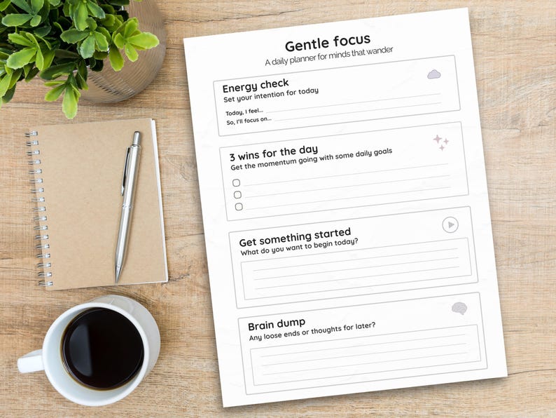 May include: A daily planner titled "Gentle Focus" with sections for daily intentions, goals, and notes. A silver pen rests on a spiral-bound notebook. A cup of coffee and a small plant are also on the wooden surface.